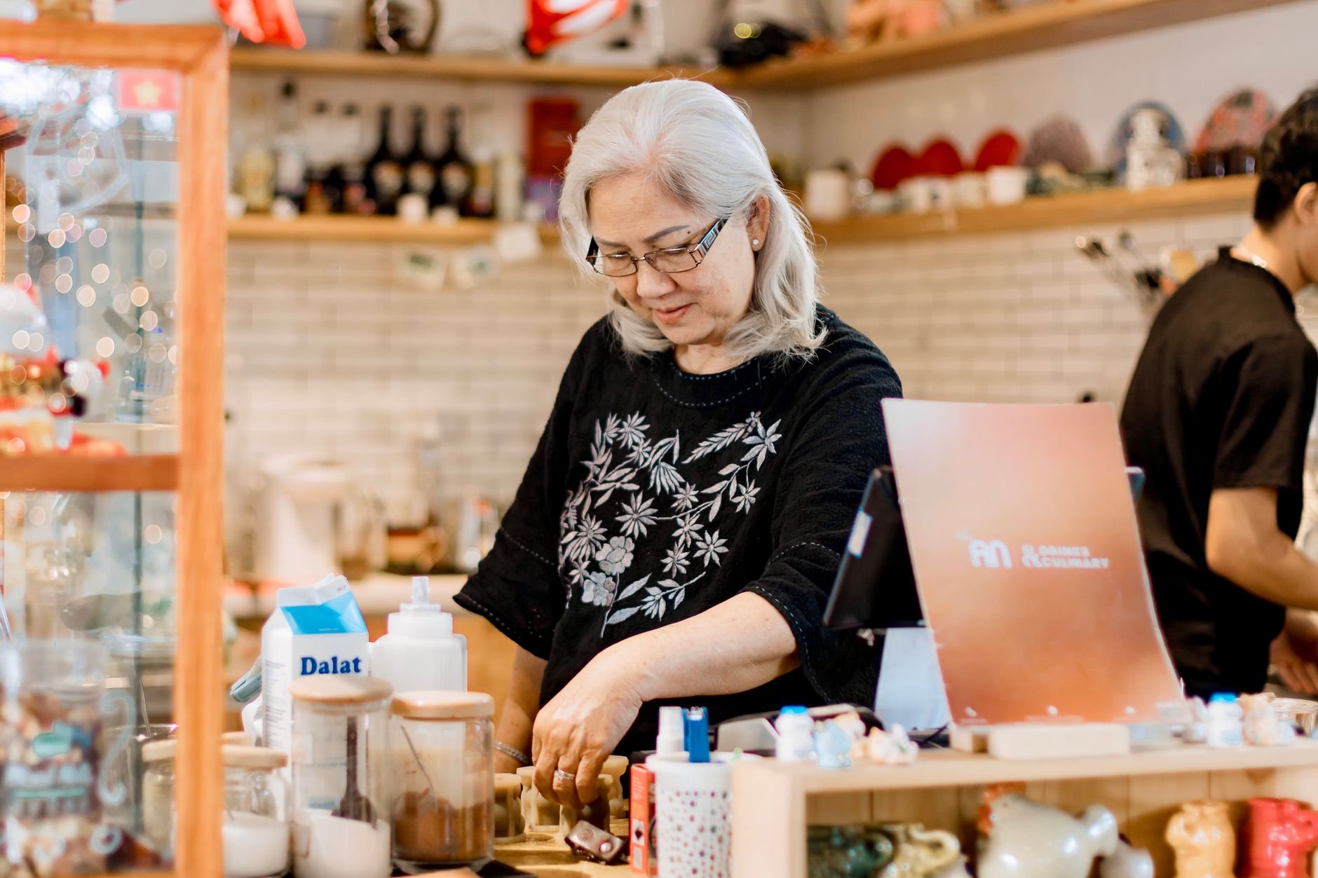 Senior woman working behind the counter in a cozy, warmly lit coffee shop.