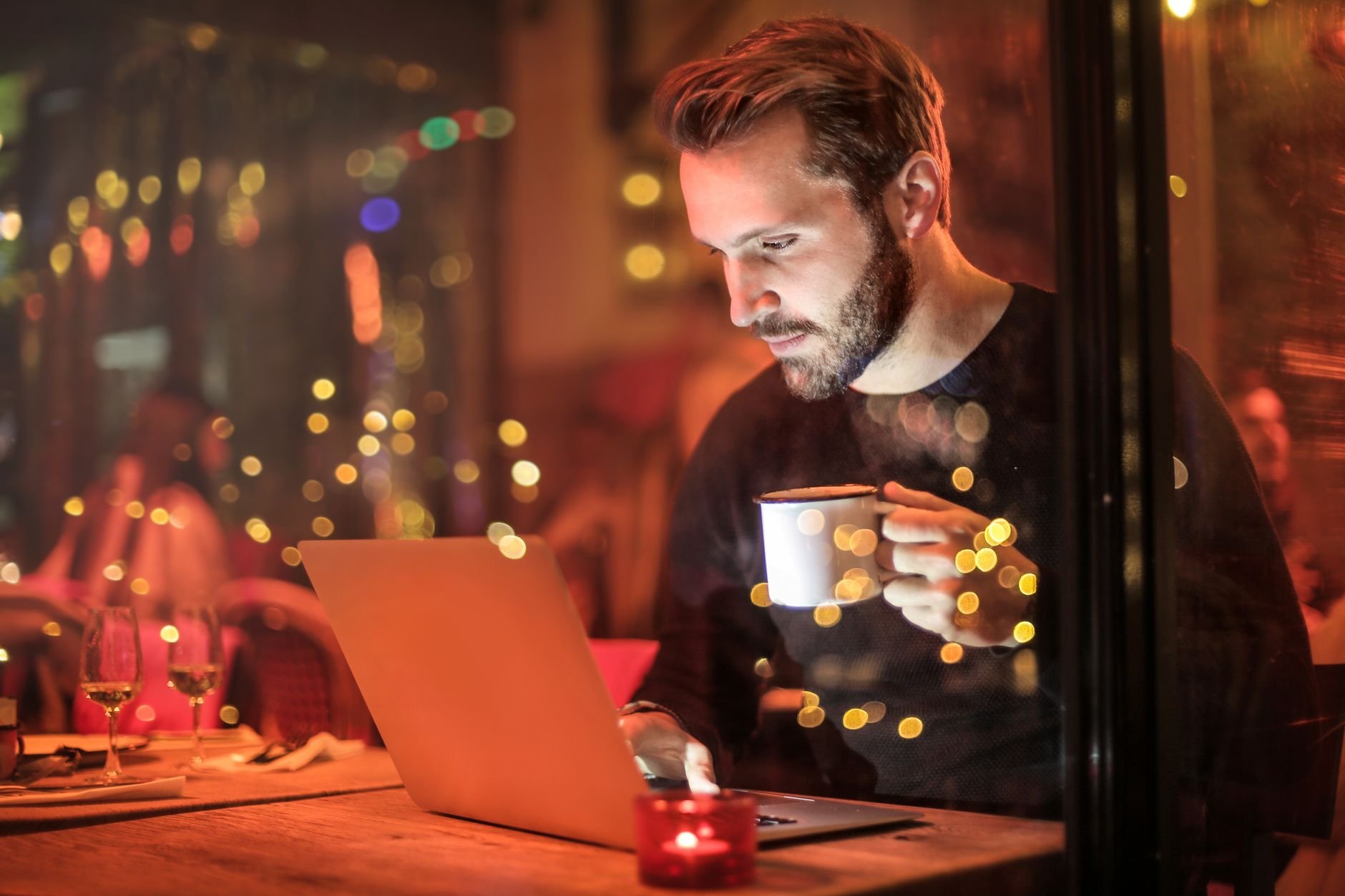 A young man with a beard working on a laptop at a cozy cafe with warm lighting.